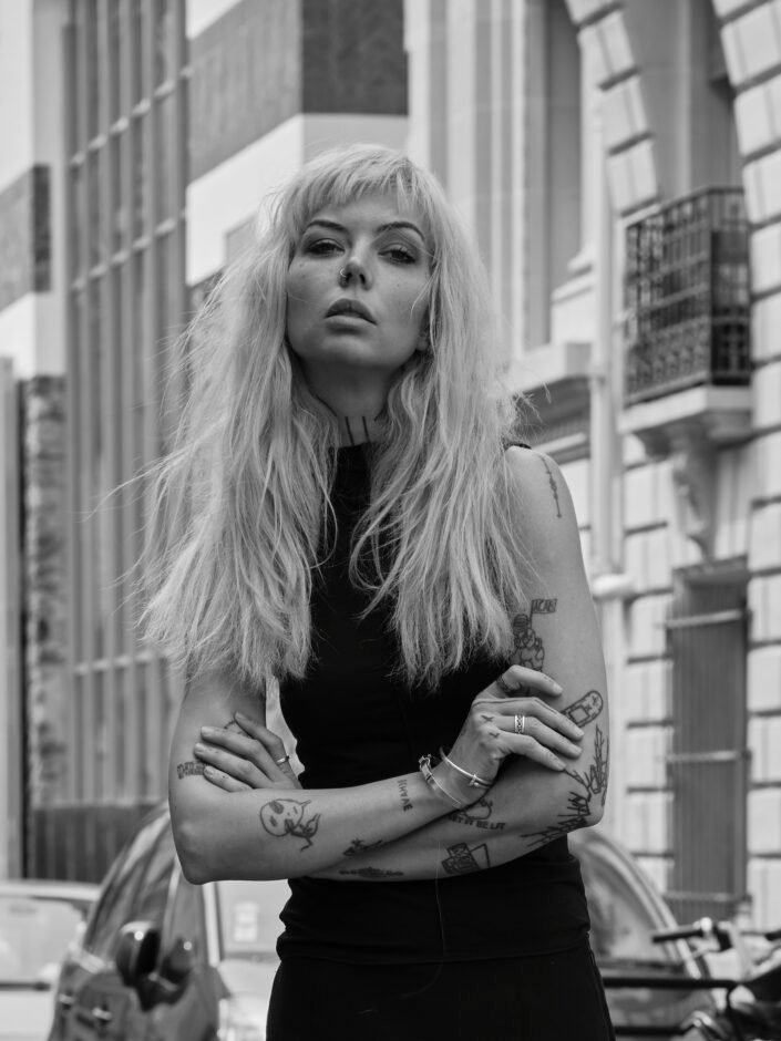 Portrait de Shai mettant en valeur ses tatouages, dans une rue parisienne